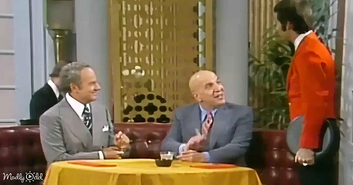 Two men in suits sit at a yellow table, engaged in conversation, while another man in an orange suit stands nearby.
