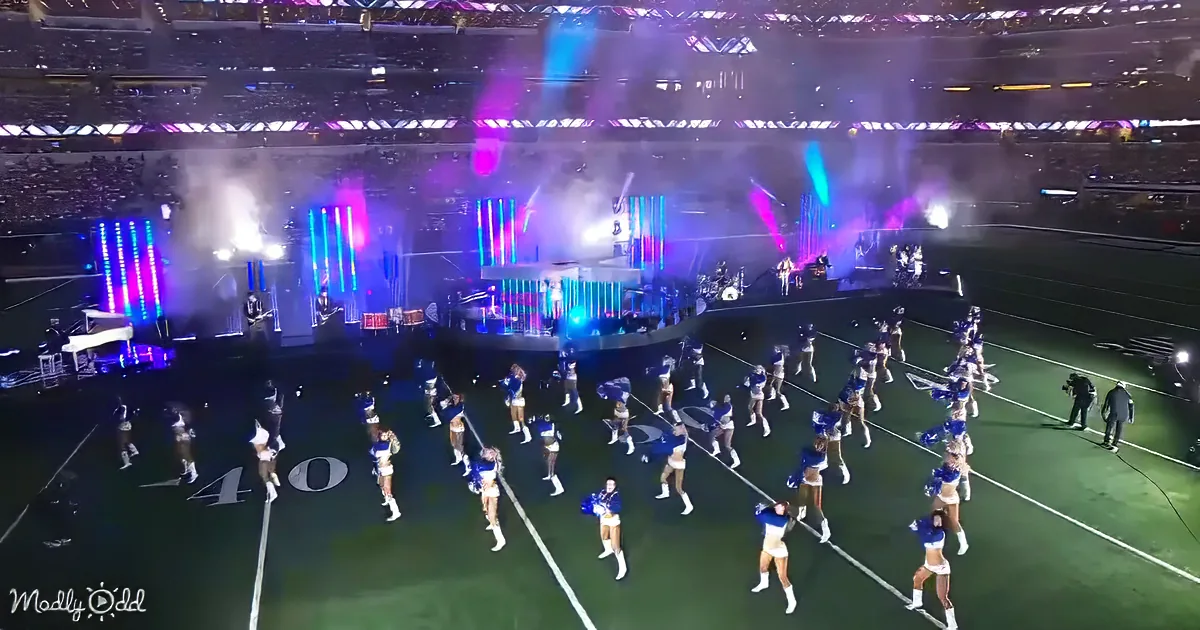Cheerleaders perform on a football field with a stage and colorful lights in the background.