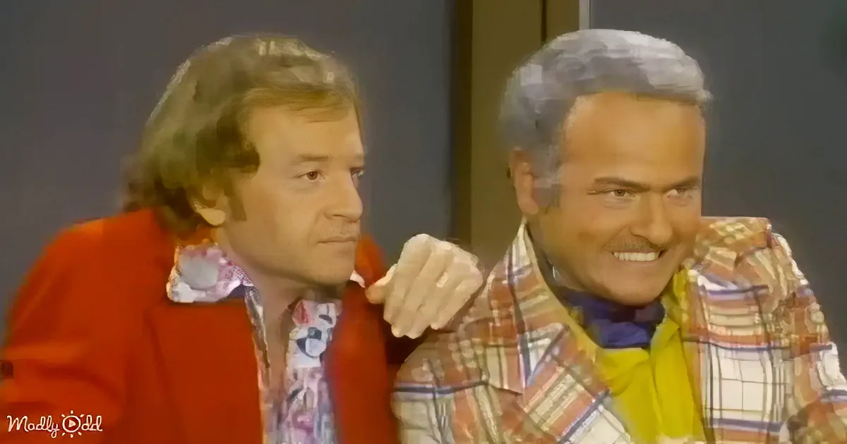 Two men in colorful attire, one with arm around the other, appear to be in a casual, possibly humorous interaction.