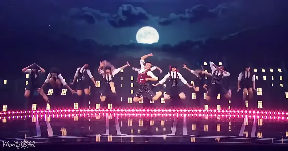 A group of dancers in coordinated outfits perform a synchronized routine on a stage with a cityscape backdrop and a full...