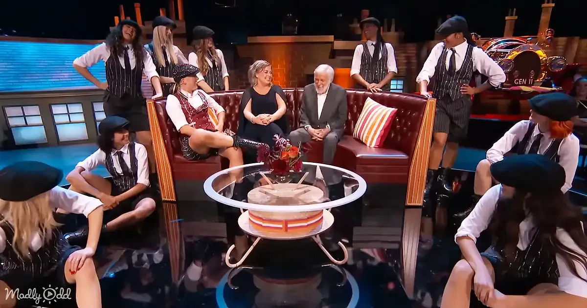 A group of people in vintage attire sits on a stage, with a man in a suit and a woman in a black dress on a red leather...