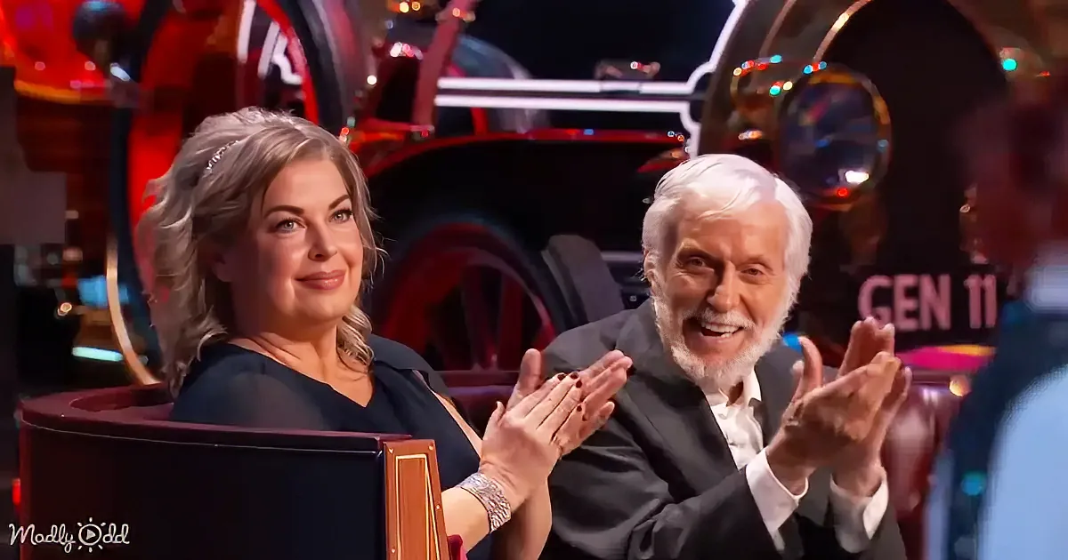 A woman and an older man are clapping, smiling, and seated in a red chair, with a colorful, illuminated background.
