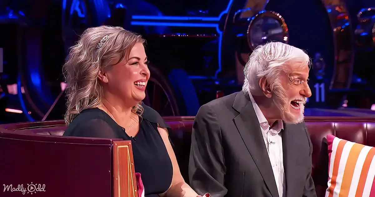 A woman and an older man are seated on a couch, both smiling and laughing, in a studio setting with a car-themed backdrop.
