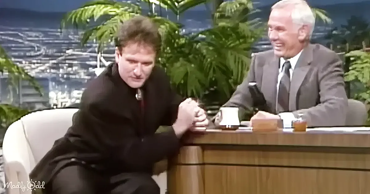 A man in a dark suit sits on a white chair, leaning forward, while another man in a suit sits at a desk with a mug, in a...