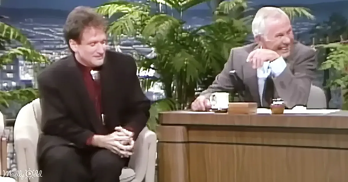 A man in a suit sits on a couch, smiling, while another man in a suit leans on a desk, also smiling, in a talk show setting.