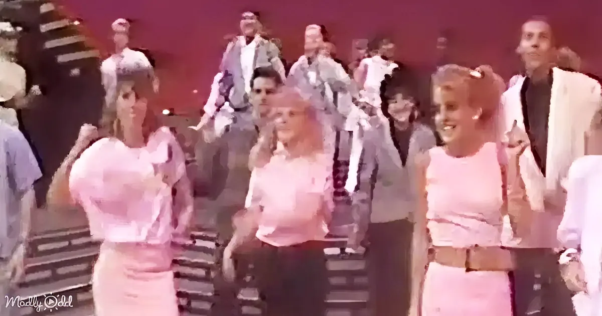 Three women in pink outfits dance energetically in the foreground, surrounded by a group of people in a lively scene.
