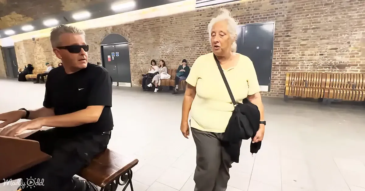 An elderly woman in a yellow shirt walks through a station, holding an umbrella, while a man in a black shirt sits on a...