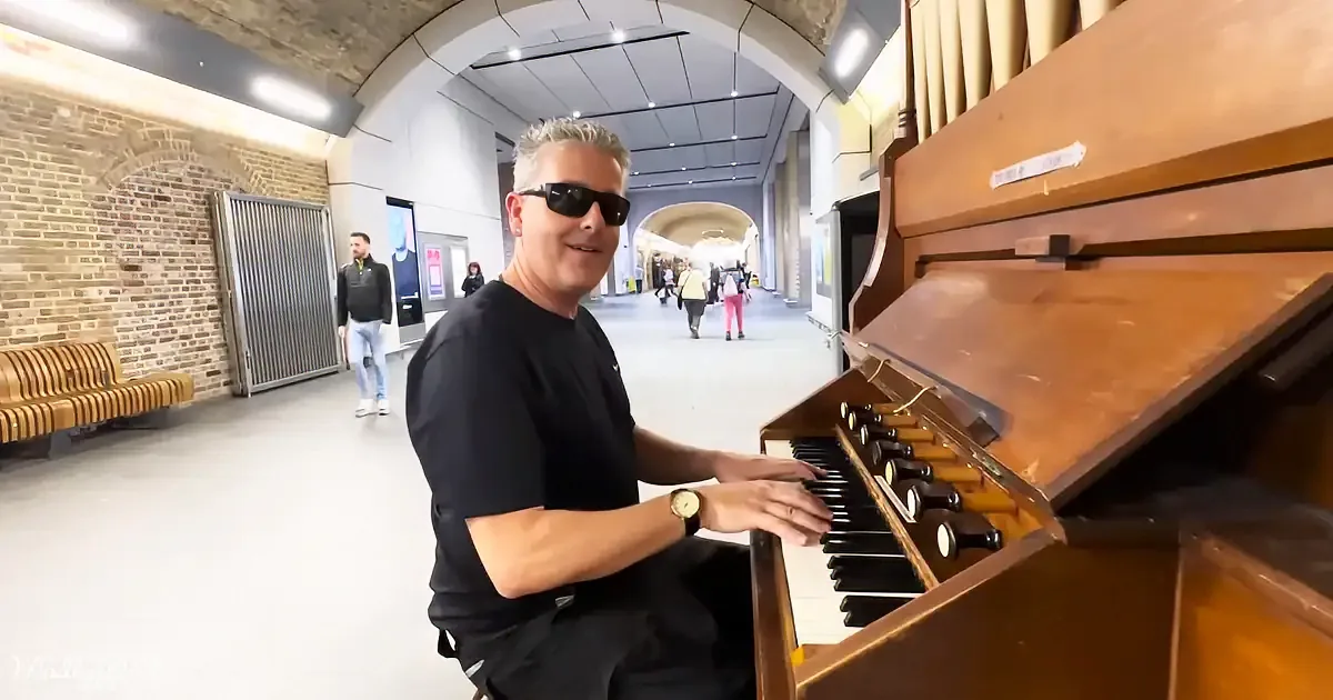 A man in black plays an organ in a spacious, well-lit indoor area with brick walls and people walking in the background.