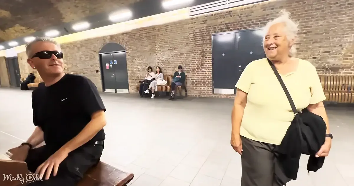 A man in sunglasses sits on a bench, while a woman in a yellow shirt stands nearby, smiling in a spacious indoor area...