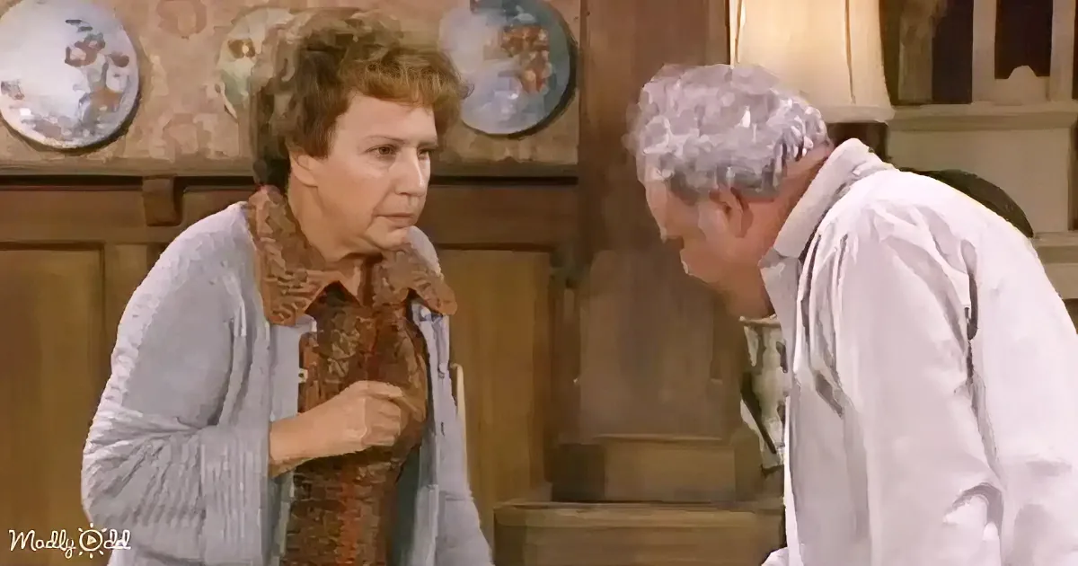 An older woman in a kitchen, holding a spoon, looks at another person with a concerned expression.