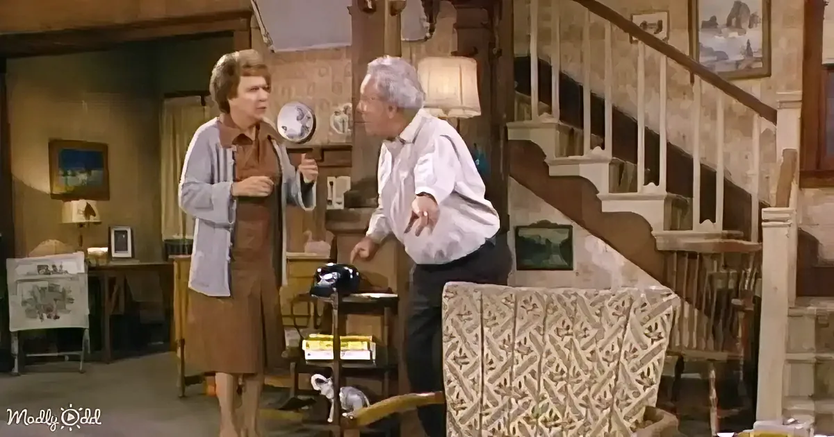 A woman in a brown dress and white cardigan stands facing a man in a white shirt and dark pants in a living room.
