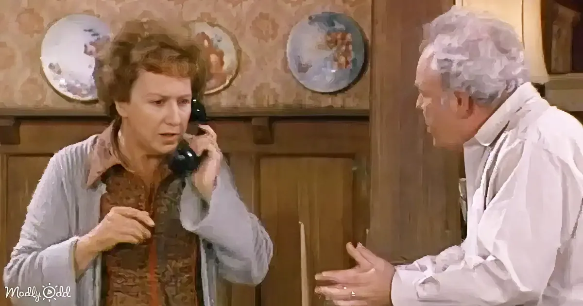 A woman holding a phone to her ear, talking, while an older man gestures beside her in a room with wooden cabinets and...