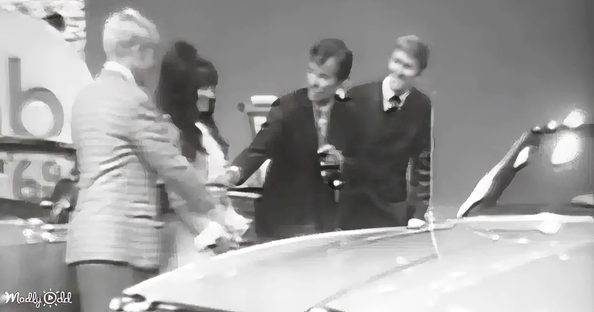 A man in a suit shakes hands with a woman in a dress near a car, with two other individuals observing in the background.
