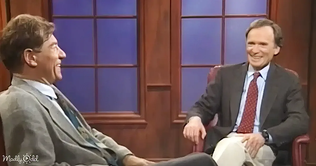 Two men in suits are seated, smiling and engaged in conversation on a talk show set.