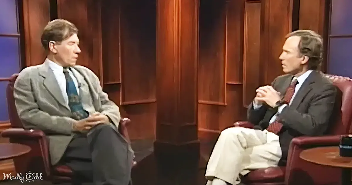 Two men in suits sit in chairs, one with hands clasped, in a studio setting with wooden paneling.
