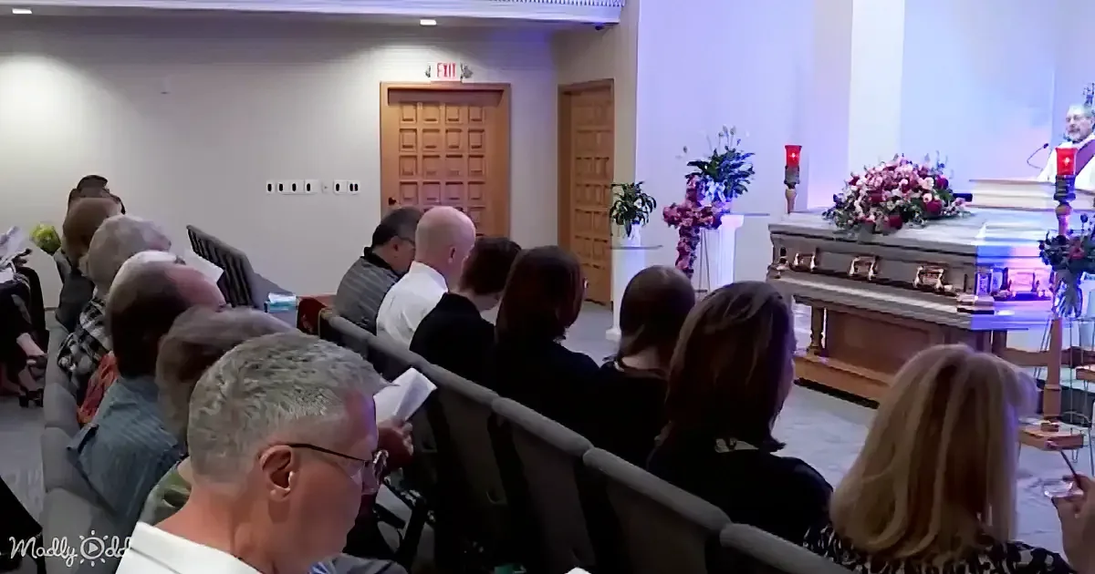 A congregation listens to a speaker at a church service with floral arrangements and a podium.