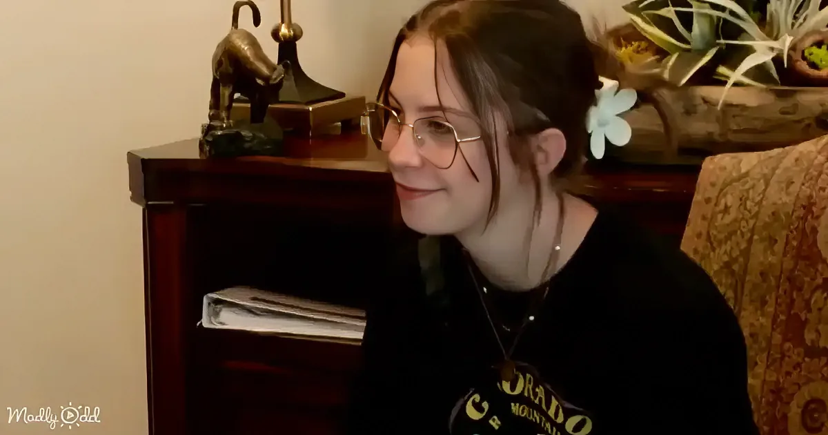 A person wearing glasses and a black shirt sits in a cozy room with a wooden cabinet, a lamp, and decorative items.