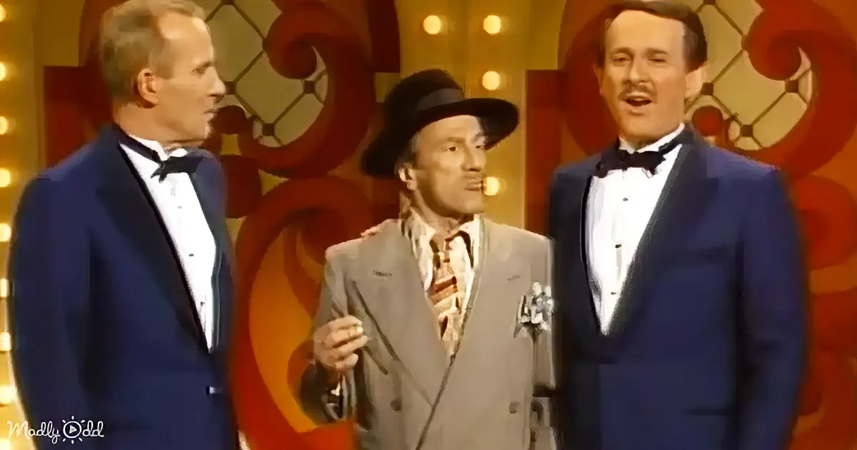 Three men in formal attire, one in a hat, stand on a stage with colorful background, possibly from a vintage show.