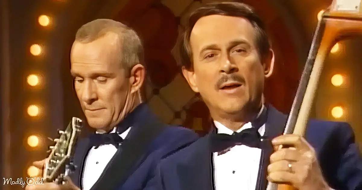 Two men in tuxedos play guitars on a stage with lights behind them.