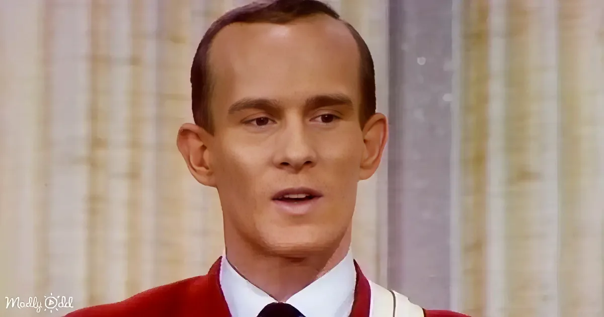 A man in a red sweater and white shirt is speaking, with a blurred background of vertical stripes.