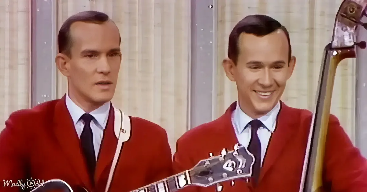 Two identical men in red suits play guitar and bass, both smiling, against a striped backdrop.
