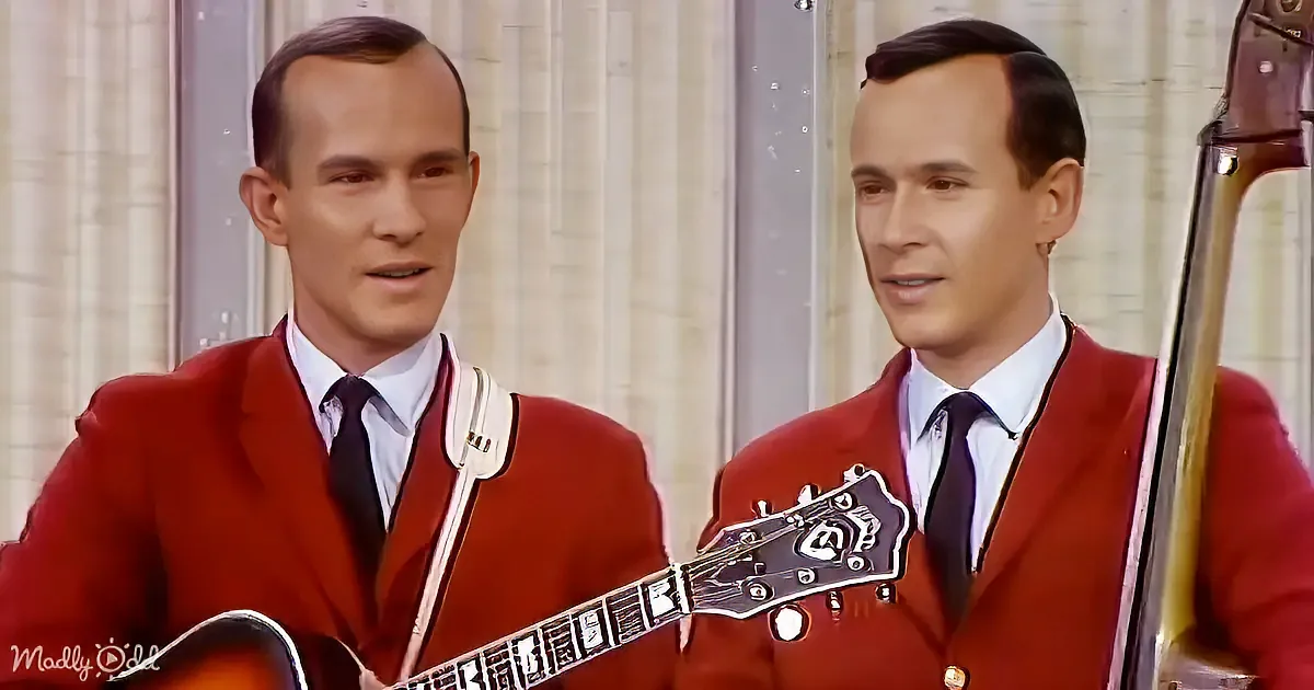 Two identical men in red suits play guitars, one with a mandolin, against a curtain backdrop.