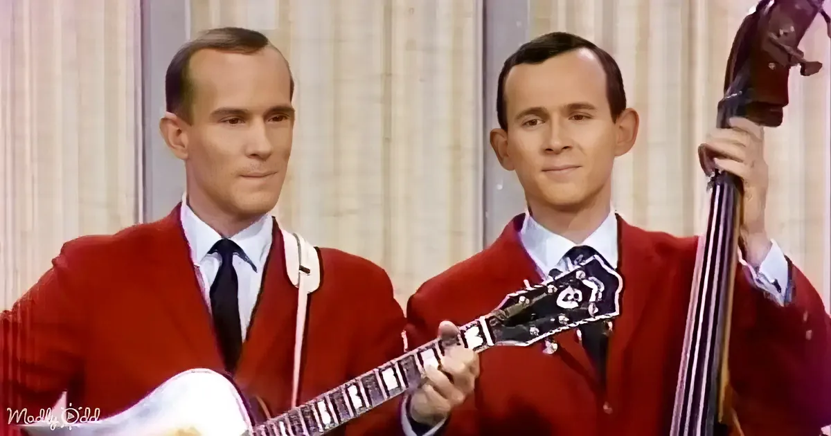 Two identical men in red jackets play guitar and bass, both with serious expressions.