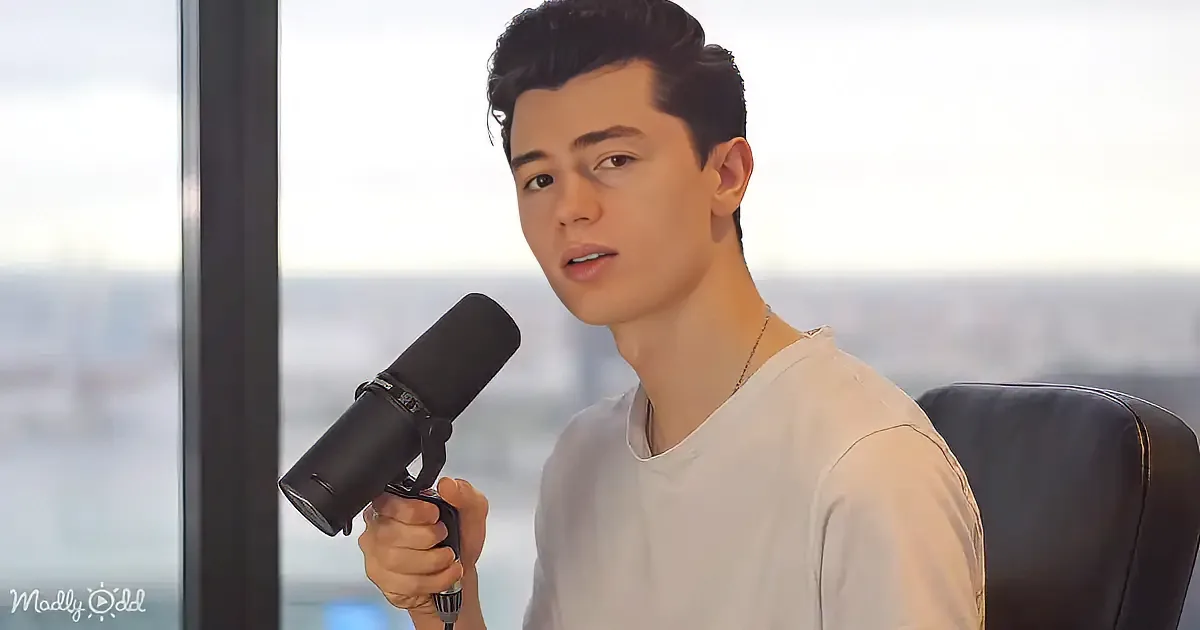 A person in a white shirt holds a microphone, seated in front of a window with a cityscape view.