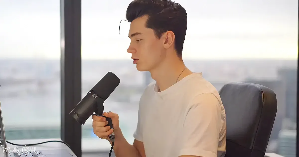 A person in a white shirt holds a microphone, seated in front of a laptop with a cityscape view through the window.