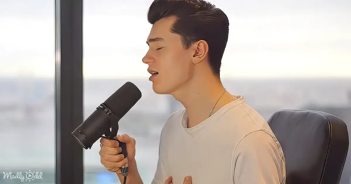 A person in a white shirt sings into a microphone, seated by a window with a cityscape view.