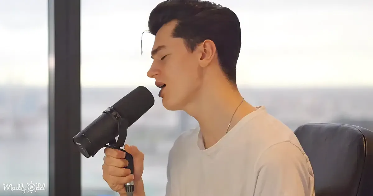 A person in a white shirt sings into a microphone, seated by a window with a cityscape view.