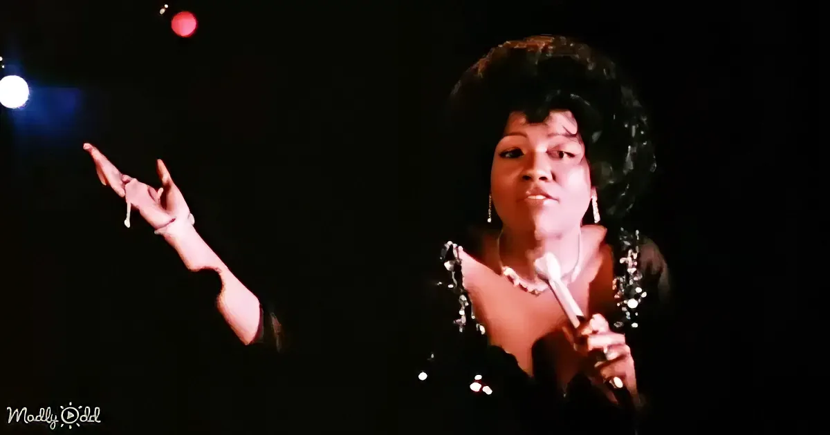 A woman in a black outfit with a microphone, gesturing with her right hand, and a red light in the background.