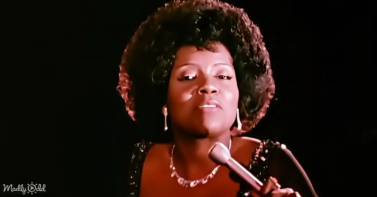 A woman with curly hair, wearing a black sequined dress and jewelry, sings into a microphone against a dark background.