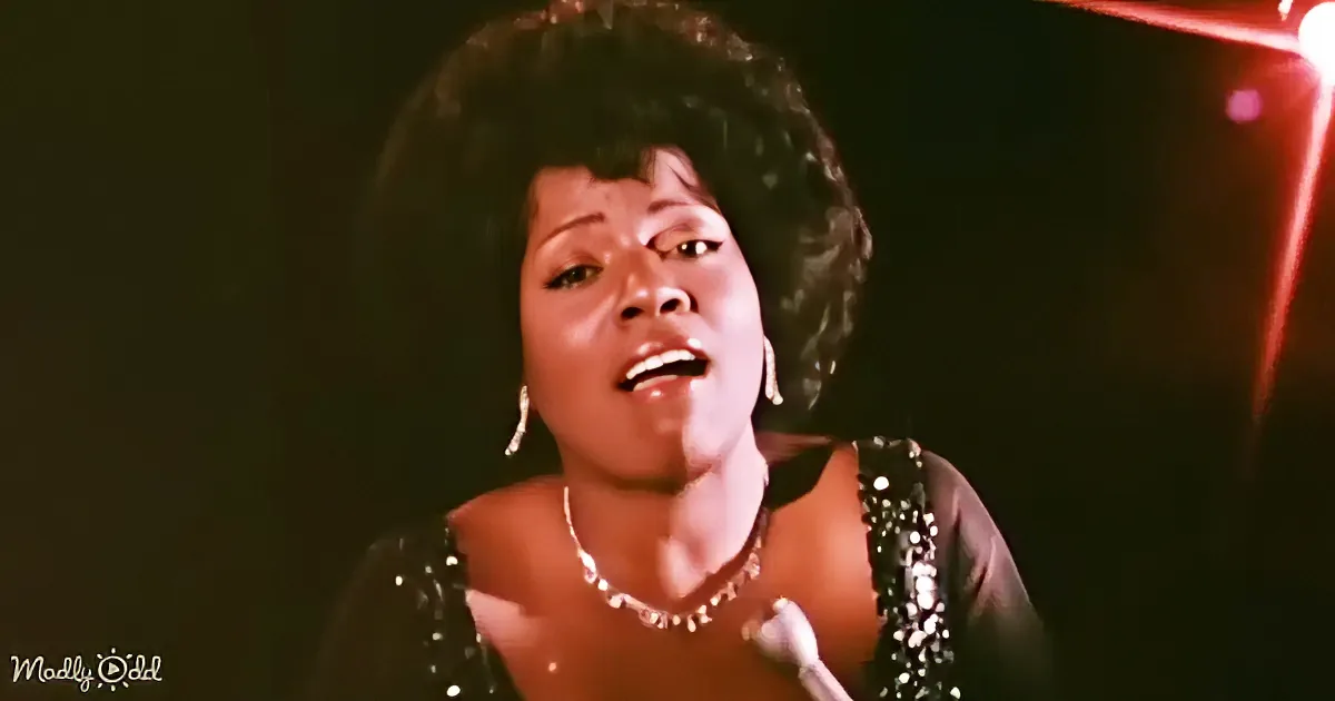A woman in a sequined dress sings passionately, wearing a necklace and earrings, with a dark background and red light.