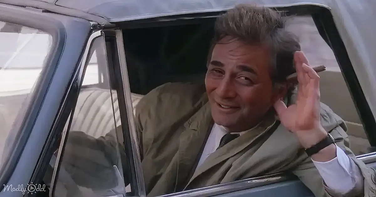 A man in a car, wearing a green jacket and white shirt, waves with his right hand while smiling.