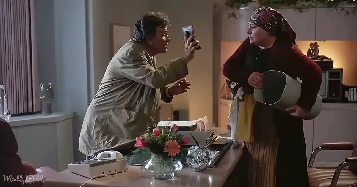 A man in a green jacket is holding a phone, while a woman in a headscarf and brown coat holds a bucket, both in a room...