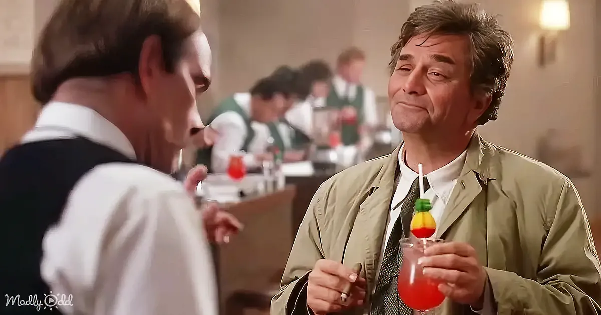 A man in a green jacket holds a drink with a colorful umbrella, looking at another man in a white shirt.