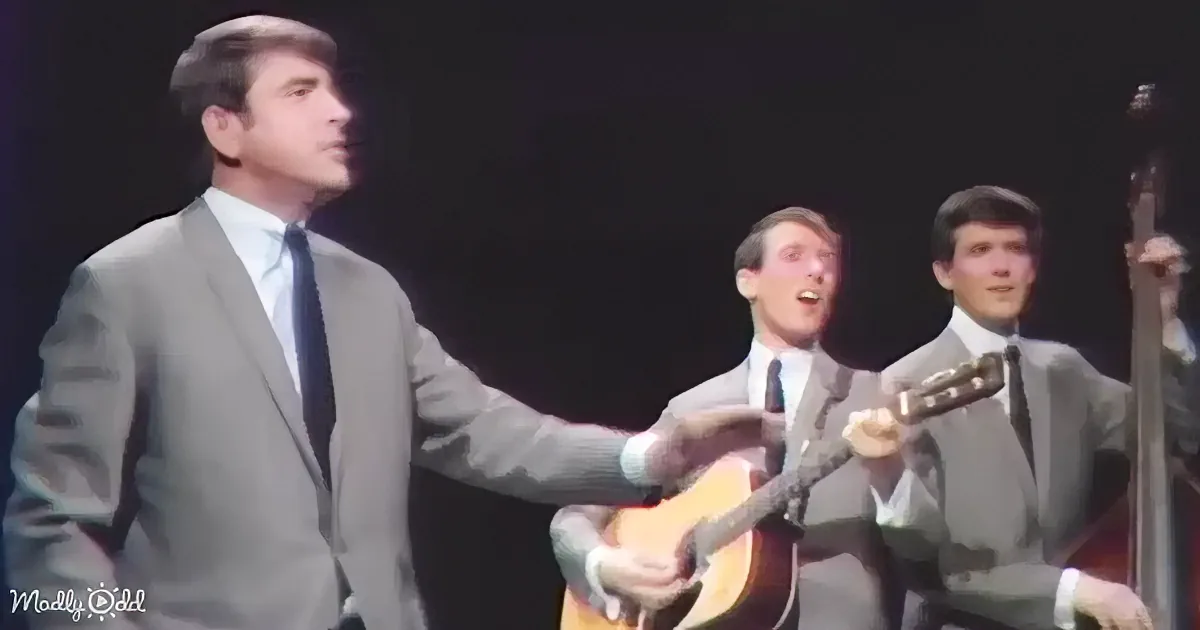 A man in a suit sings passionately, gesturing with his right hand while another man plays guitar in the background.