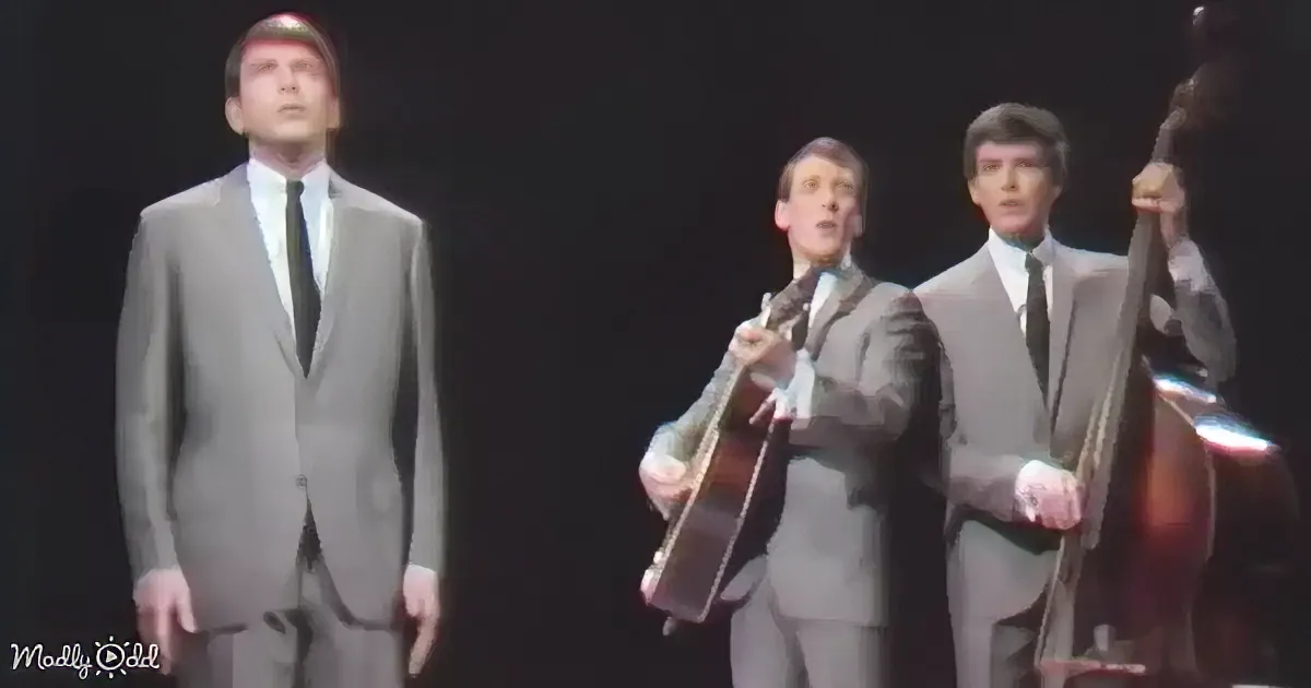 A man in a suit sings on stage, accompanied by musicians playing guitar and double bass.