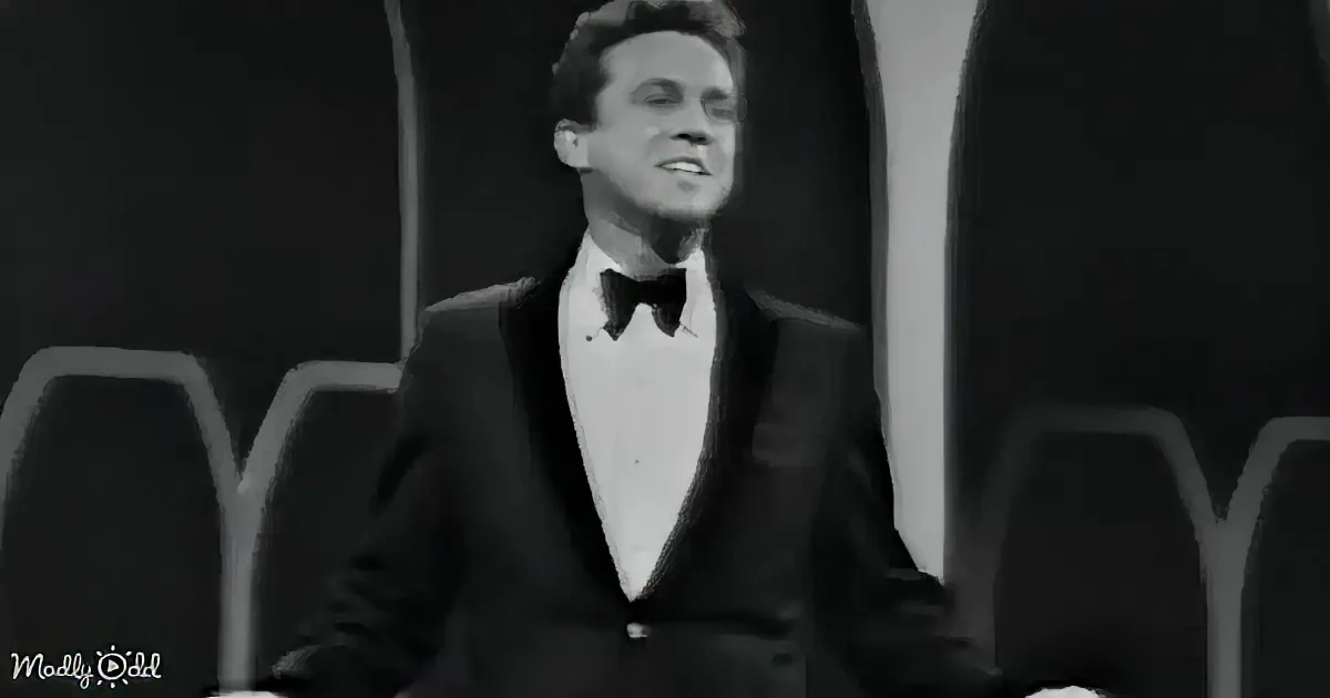 A man in a tuxedo smiles, seated on stage with a black-and-white backdrop.