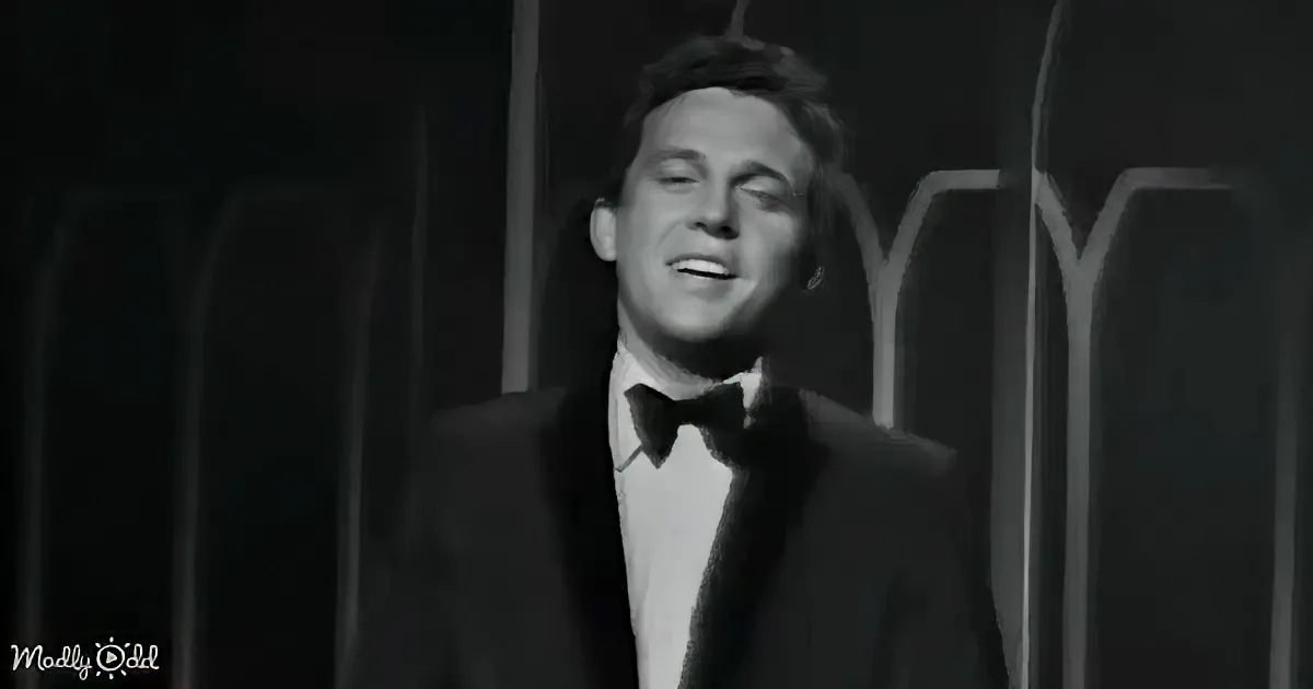 A man in a tuxedo smiles, wearing a bow tie, against a dark, patterned background.