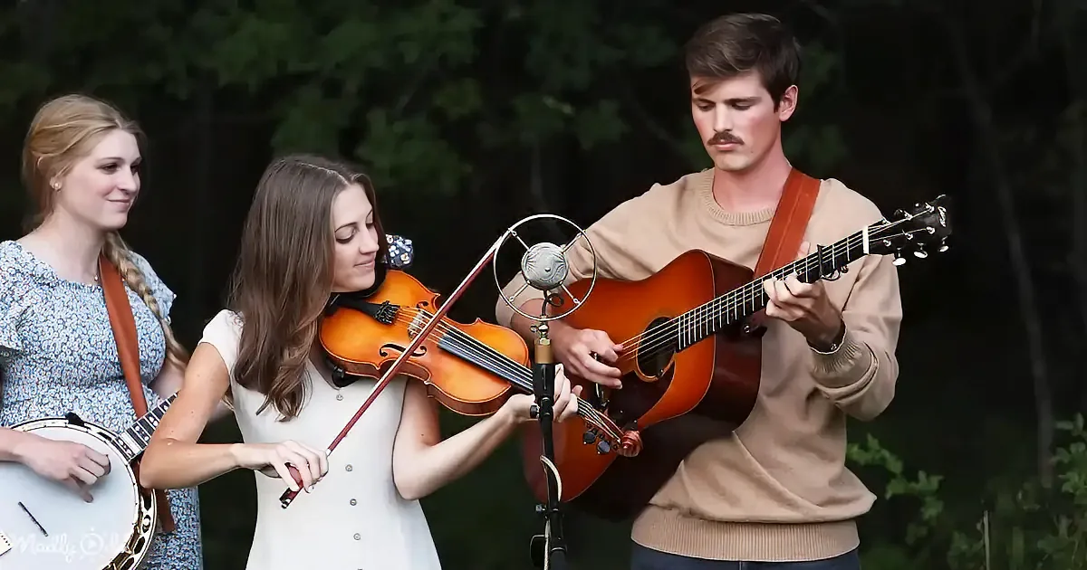A man plays guitar while a woman in white holds a violin, another woman plays banjo nearby.