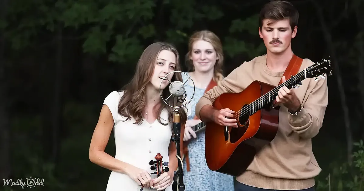 A woman sings into a microphone while a man plays guitar, with another woman in the background.