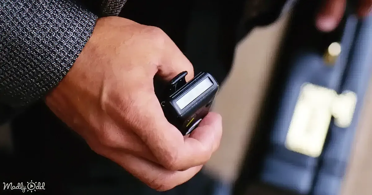 A hand holds a small black device with a screen and buttons, possibly a pager or remote control.