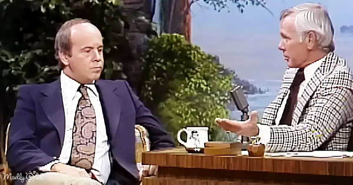 A man in a suit sits at a table with a coffee mug, engaged in conversation with another man gesturing.