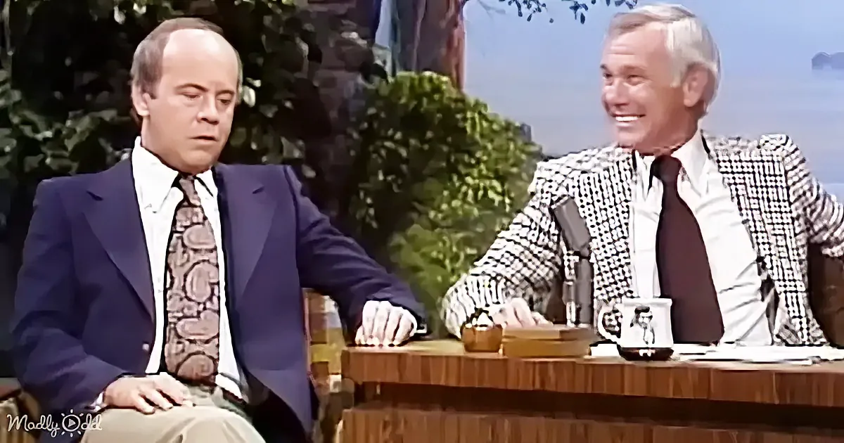 Two men in suits sit at a desk, one smiling, the other looking down, with a mug and papers on the desk.