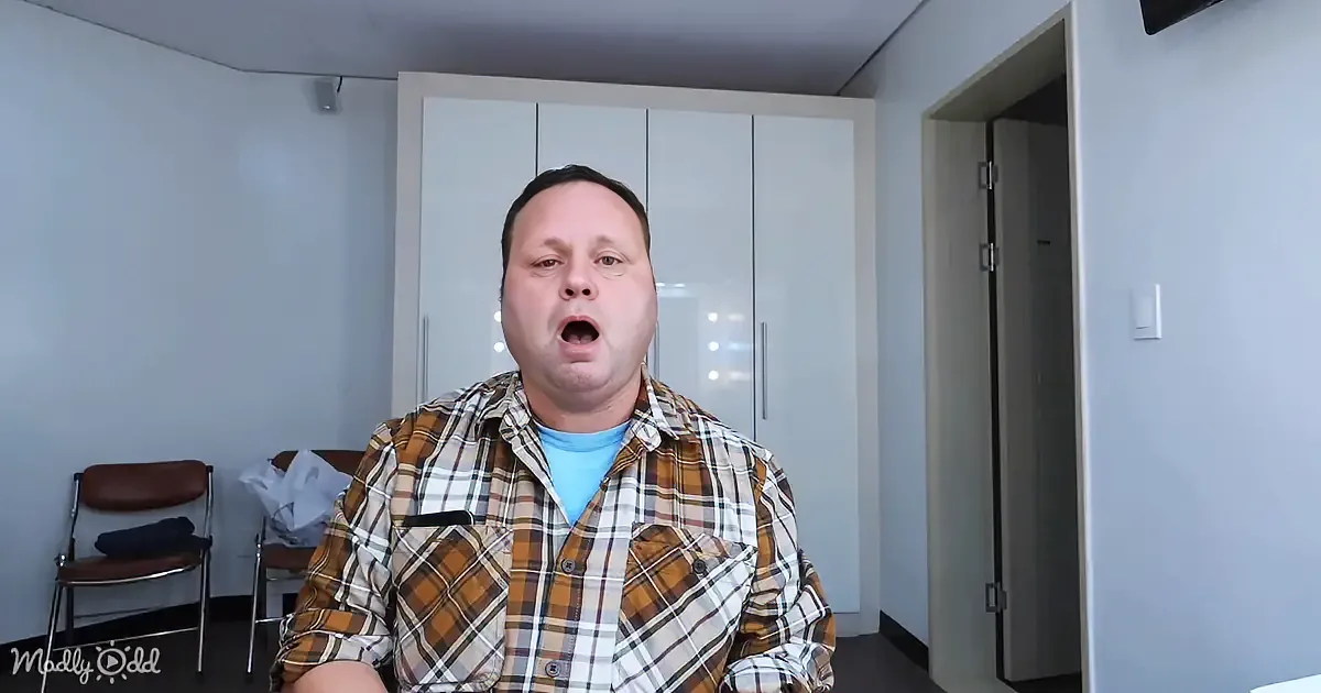 A man in a plaid shirt stands in a room with a closet and chairs, mouth open.