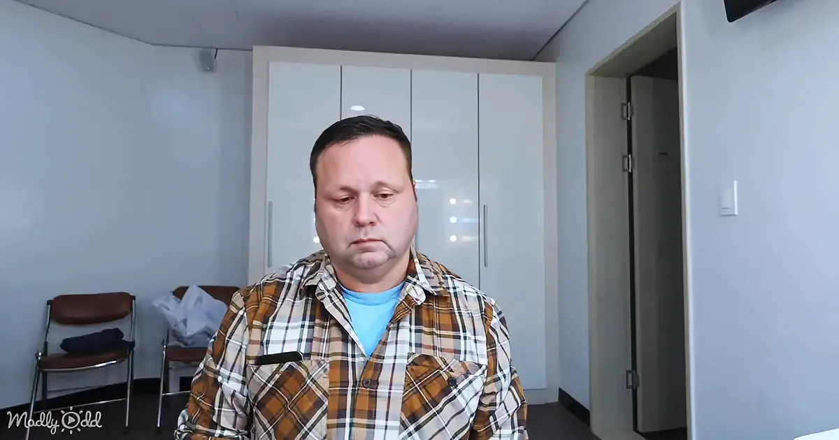 A man in a plaid shirt stands indoors, looking downward, with a chair and closet in the background.