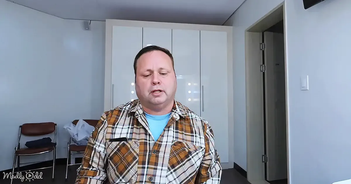 A man in a plaid shirt stands indoors, looking slightly downward, with a chair and white cabinets in the background.