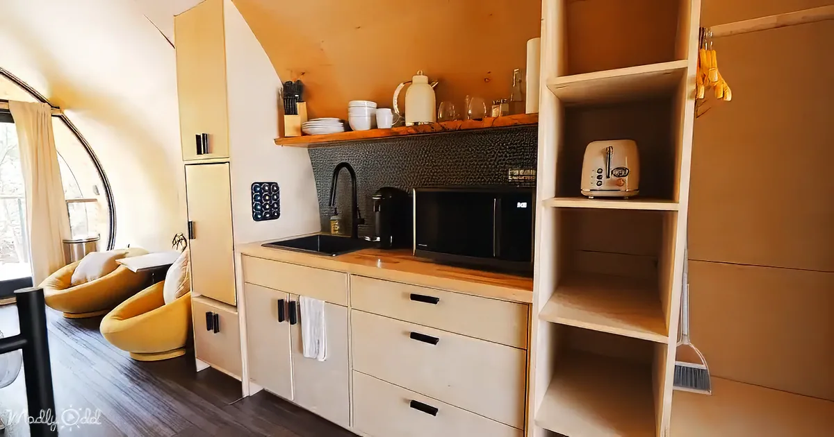 Compact kitchen with wooden shelves, microwave, sink, and yellow chairs in a cozy space.